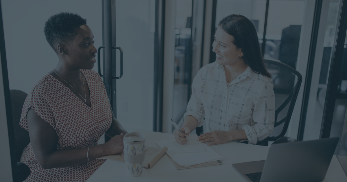 Two women in a modern office having a conversation or meeting. One woman is taking notes in a notebook.
